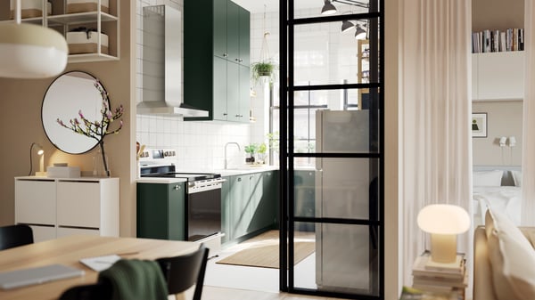 An open kitchen with deep green HAVSTORP kitchen fronts and wall cabinets up to the ceiling, next to a bright living room.