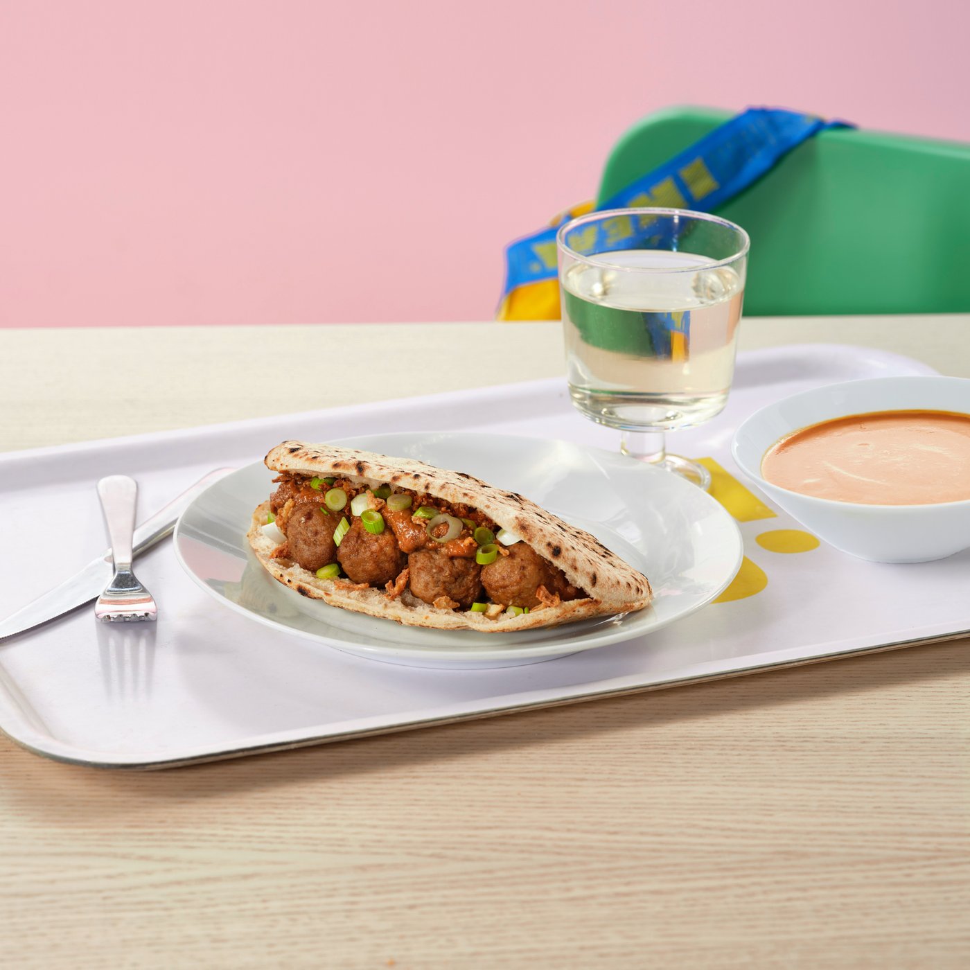 A pita bread filled with meatballs, spring onion, and peanut sauce on an IKEA tray, served with a bowl of tomato soup and a soft drink.