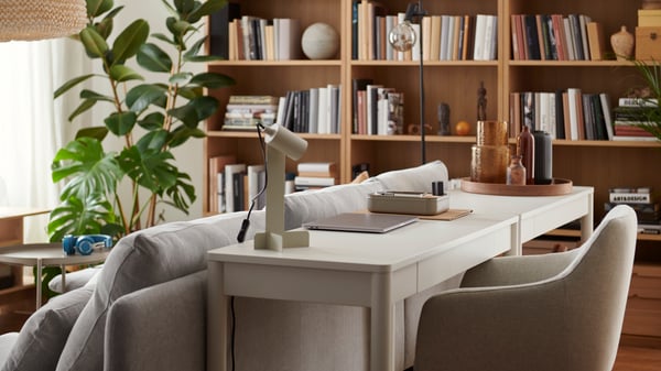 A living space with a small workstation placed behind a sofa, consisting of a white TONSTAD desk and a FLOTTILJ lamp.