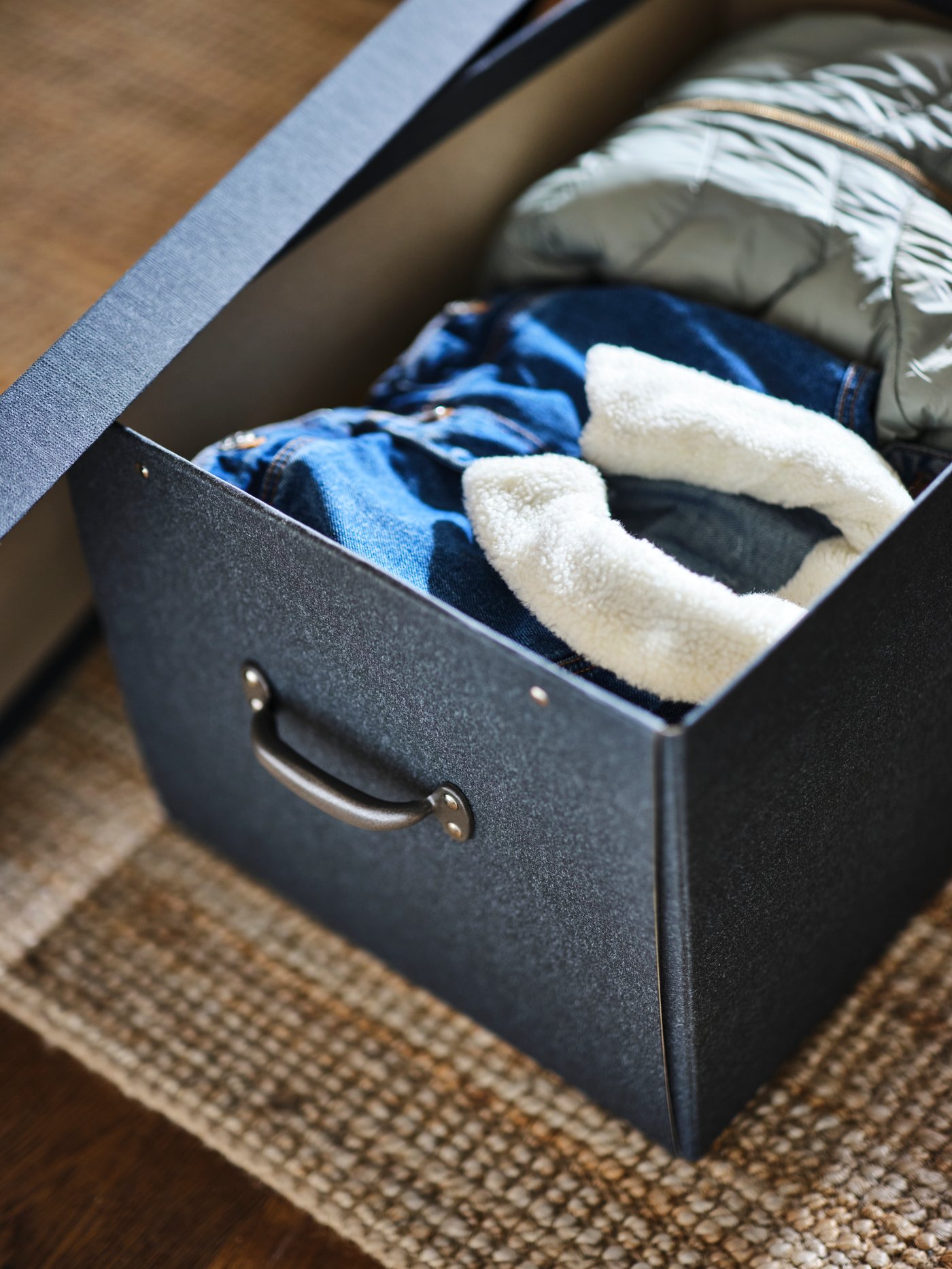 An open dark grey TJOG storage box with lid on a rug with folded clothes inside.