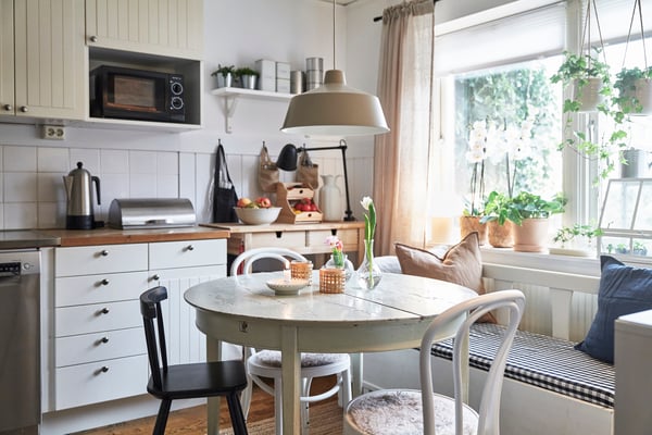 En bild på ett ljust kök med ett vitt runt matbord med två vita köksstolar, en svart köksstol och en soffa som står under ett fönster. Ovanför bordet hänger en vit lampa.