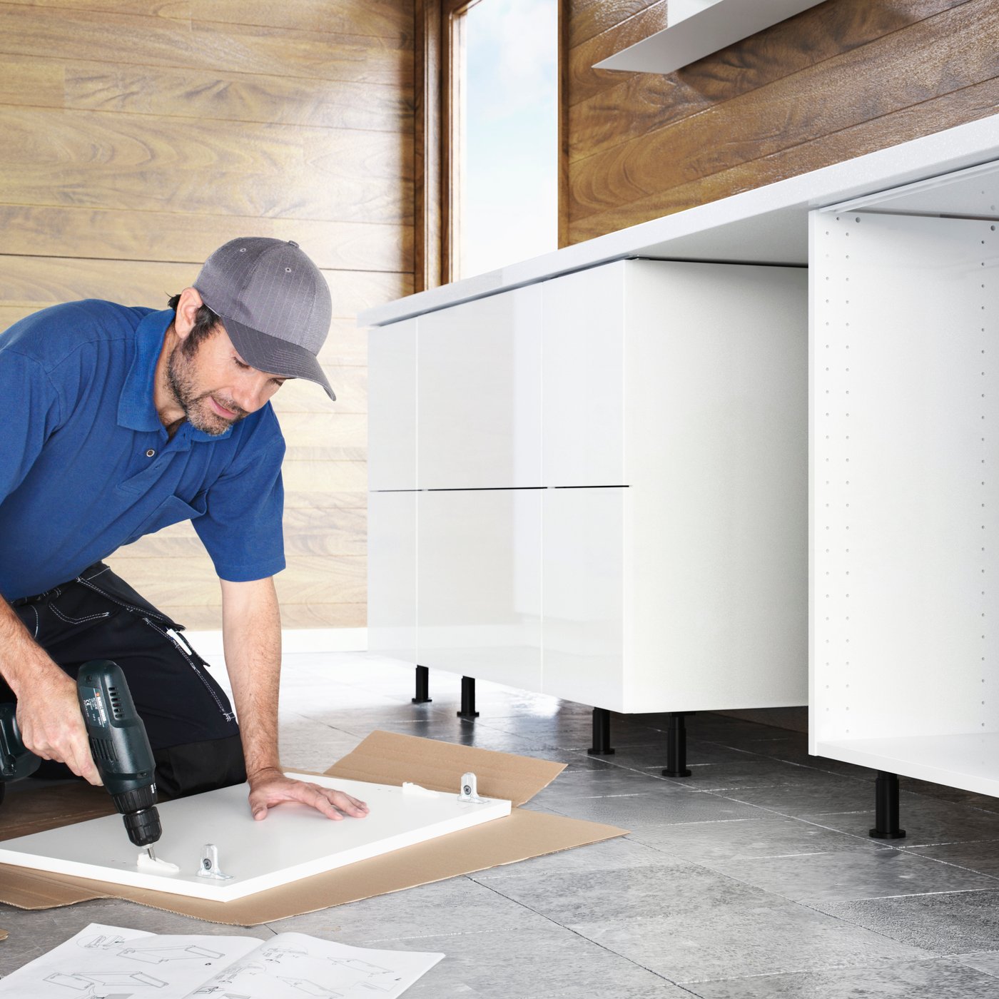 Küchenmontageservice: Eine Person in Arbeitskleidung kniet auf einem gefliesten Fußboden in einer zum Teil fertigen Küchen und bohrt Schrauben in eine weiße Front.