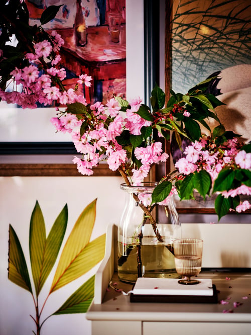 Salon avec un vase en verre blanc contenant des fleurs, un verre d'eau, des cadres, un rangement en bois et des plantes d’intérieur.
