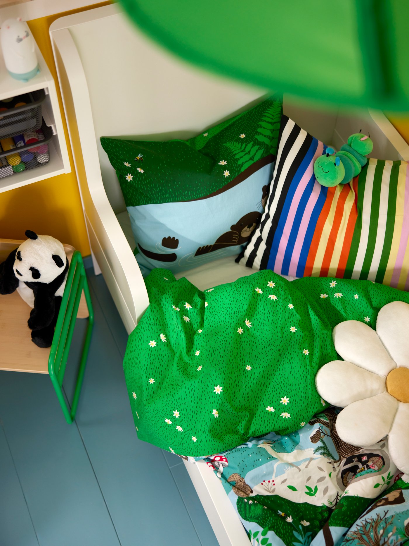 Blick von oben auf ein Kinderbett mit SKOGSDUVA Bettwäsche. Drumherum sind Kissen und Stofftiere verteilt.