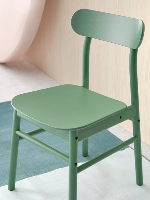 A close-up of a green RÖNNINGE chair on a rug by a wall shows its rounded seat, angled backrest and visible wood grains.