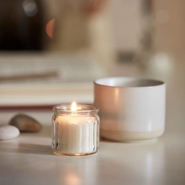 A lit Scandinavian Woods/white ADLAD scented candle in glass and a stoneware mug on a white KALLAX desk.
