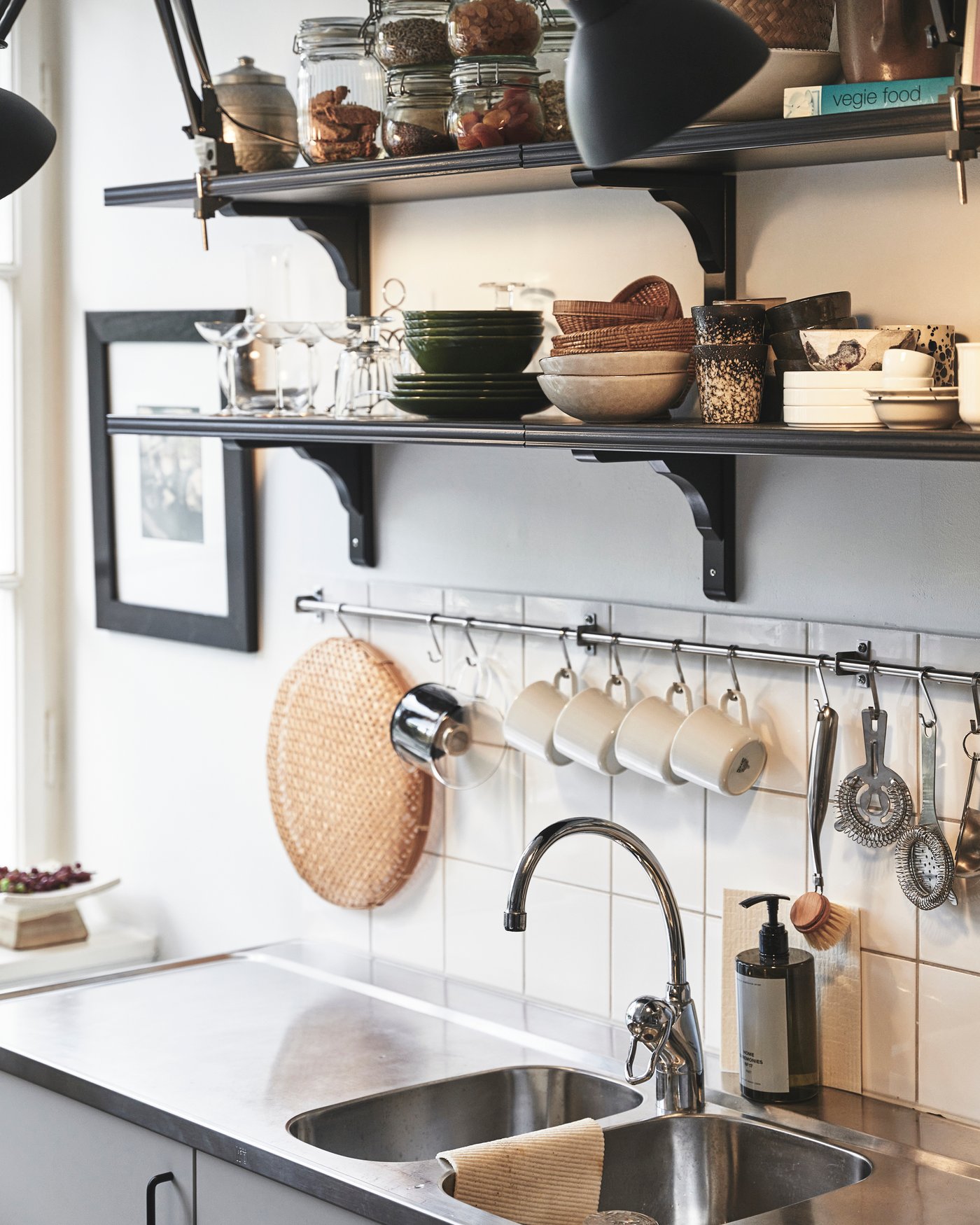 Svarta hyllor mot en vit vägg. Under hänger en rostfri köksstång där det hänger muggar och diverse köksredskap.