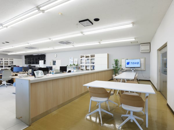 長いカウンターと白いチェアを並べた受付エリア。奥に収納棚とワークスペースが続く、シンプルなオフィスレイアウト。