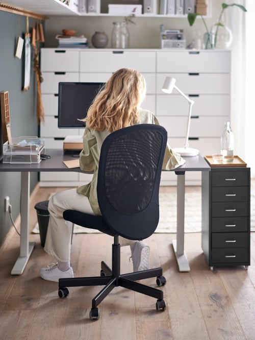 O femeie așezată pe un scaun de birou FLINTAN negru lângă un birou RODULF cu înălțime reglabilă cu un computer, o lampă de lucru și o unitate cu sertare.