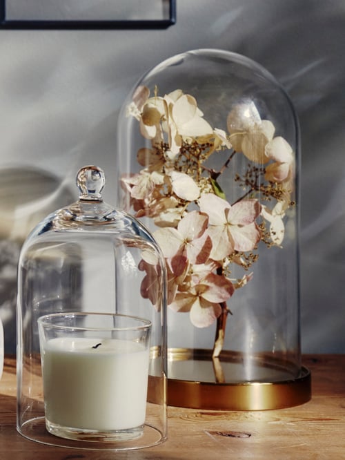 Sur une table, deux cloches en verre BEGÅVNING. Sous l'une d'elles, des fleurs séchées; sous l'autre, une bougie éteinte.
