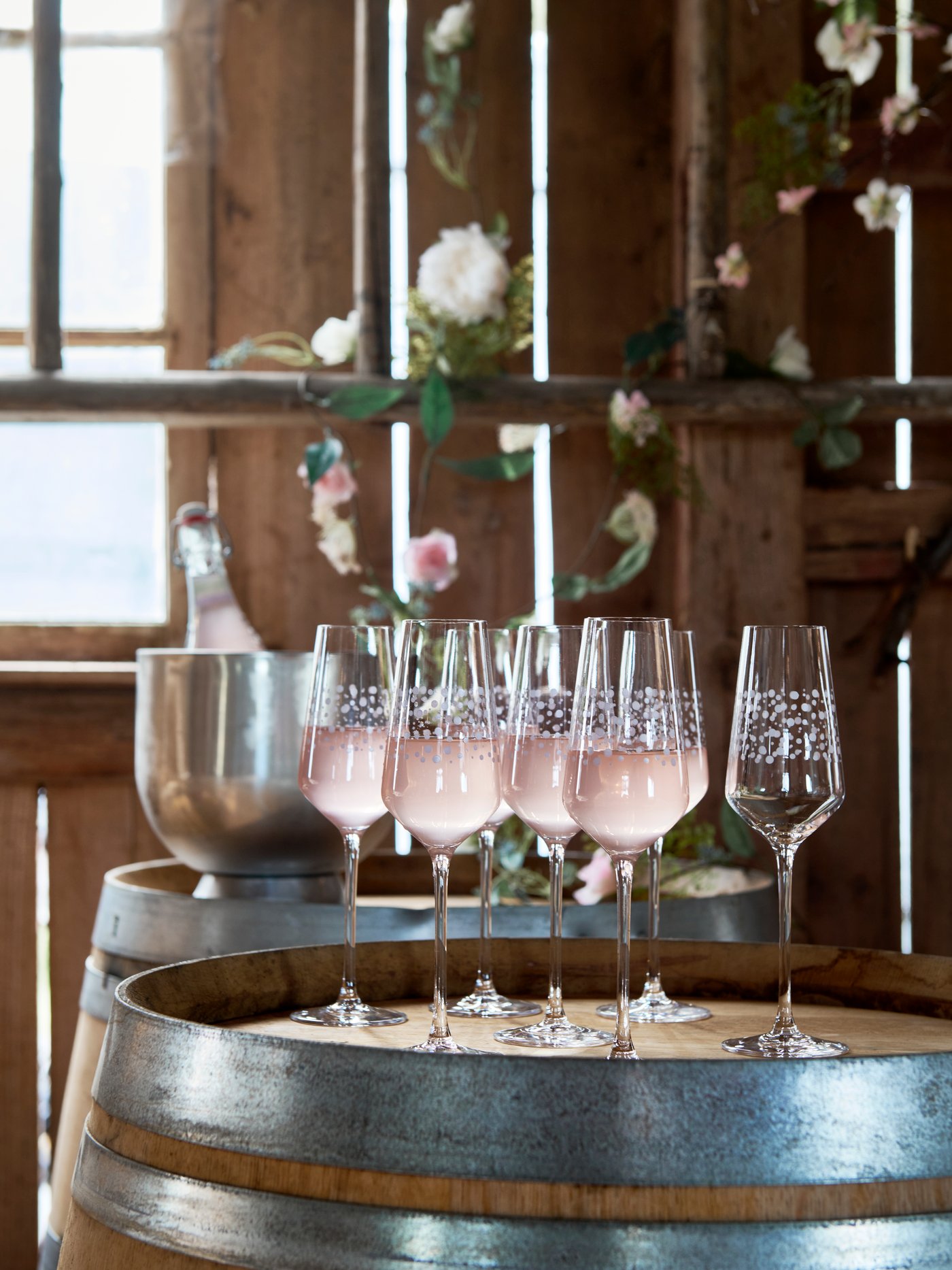 Seven INBJUDEN glasses, set on a barrel and filled with pink lemonade, await guests in a rustic party setting.