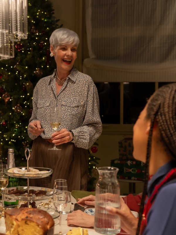 Una donna sorridente con i capelli corti e grigi è in piedi a capotavola e brinda con un bicchiere di spumante. Sullo sfondo, un albero di Natale illuminato.