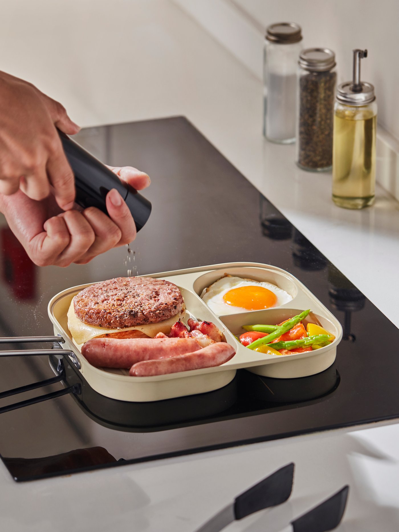コンロの上で仕切り付きフライパンに調味料をかけている様子。パンに乗ったハンバーグ、ソーセージ、目玉焼き、炒め野菜がフライパンに入っており、奥にスパイスやオイルの容器が見える。