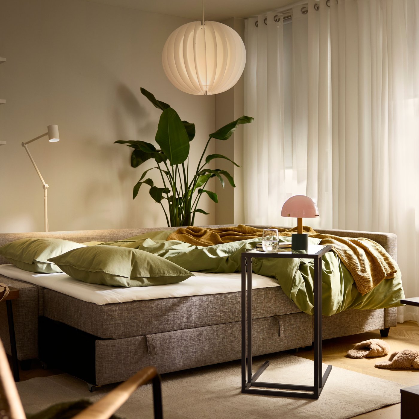 A FRIHETEN sofa bed in bed mode with its bedding undone, in a softly lit living room with curtains and blinds mostly shut.