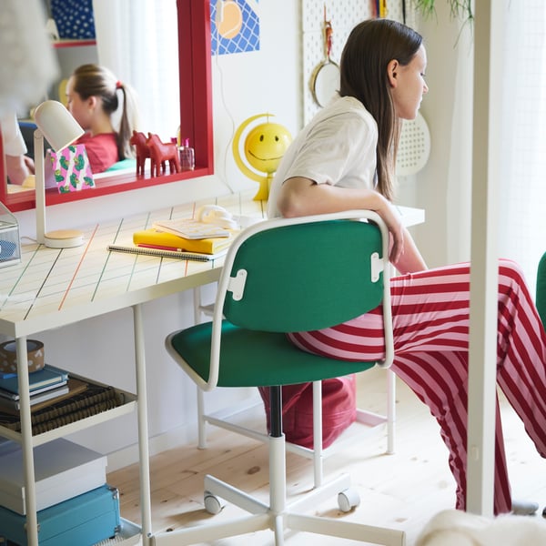 A teenager sitting on a green ÖRFJÄLL swivel chair by a desk with a red mirror and SKÅDIS pegboard in the background.