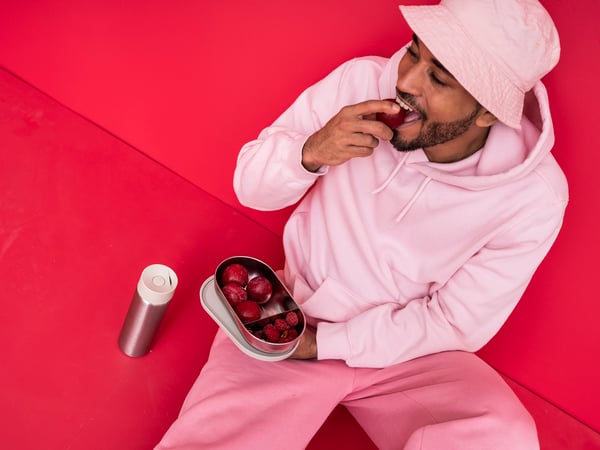 Person klädd i ljusrosa hoodie och byxor sitter mot en röd bakgrund, håller en matlåda med röda plommon och äter ett av dem. Bredvid ligger en silverfärgad termos.