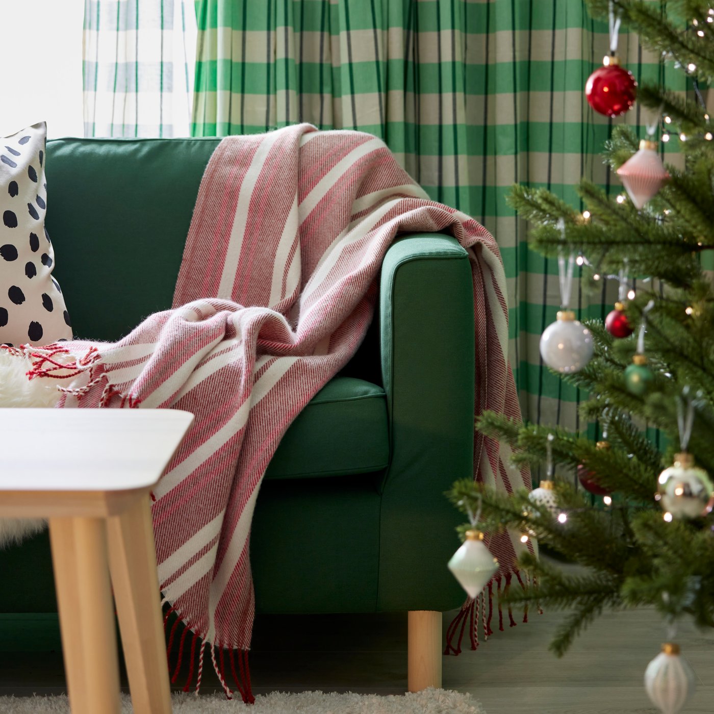 Un living con un sofá verde al costado de un parbol de navidad. Sobre el sofá una manta roja con blanco.