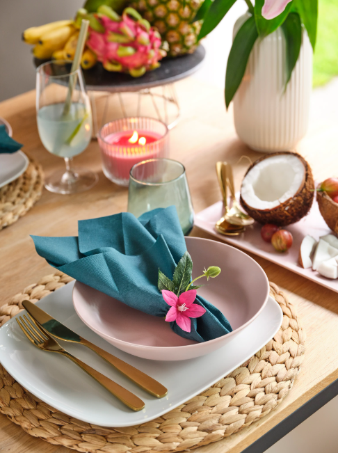 Sommerlich gedeckter Tisch mit rosa Teller, blauer Serviette und Blüte, daneben exotische Früchte, Kerze und Glas.