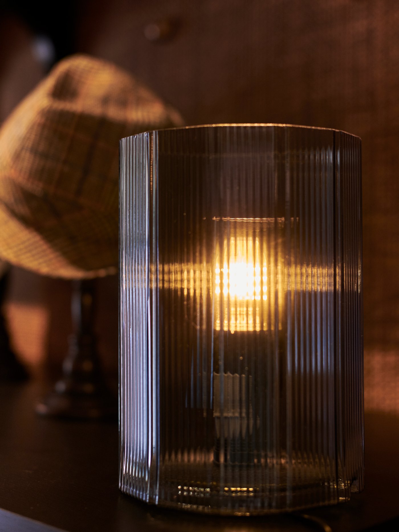 A blue MIKROKLIN glass lamp on a dark surface with a brown hat behind. The bulb is switched on and it gives off a soft glow.