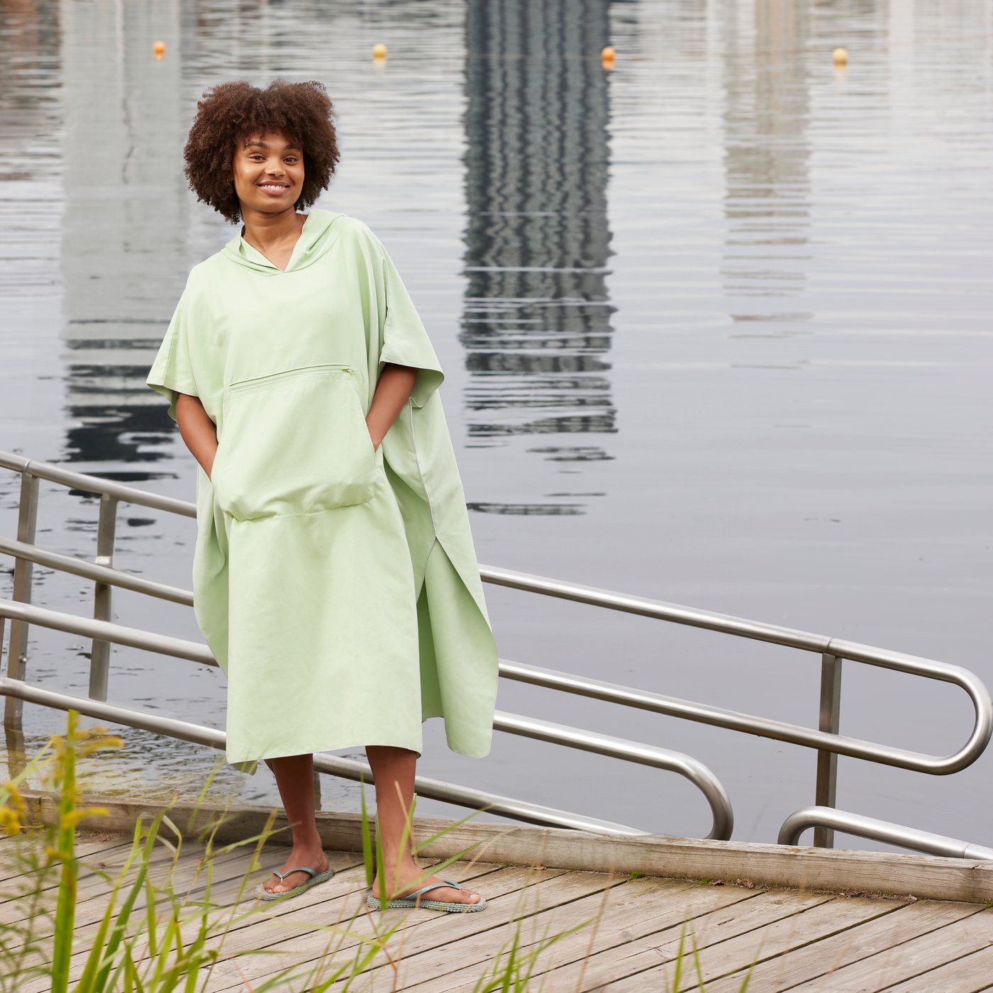A girl is standing on a dock next to a lake, wearing the DAJLIEN bath poncho with hood in light green and smiling.