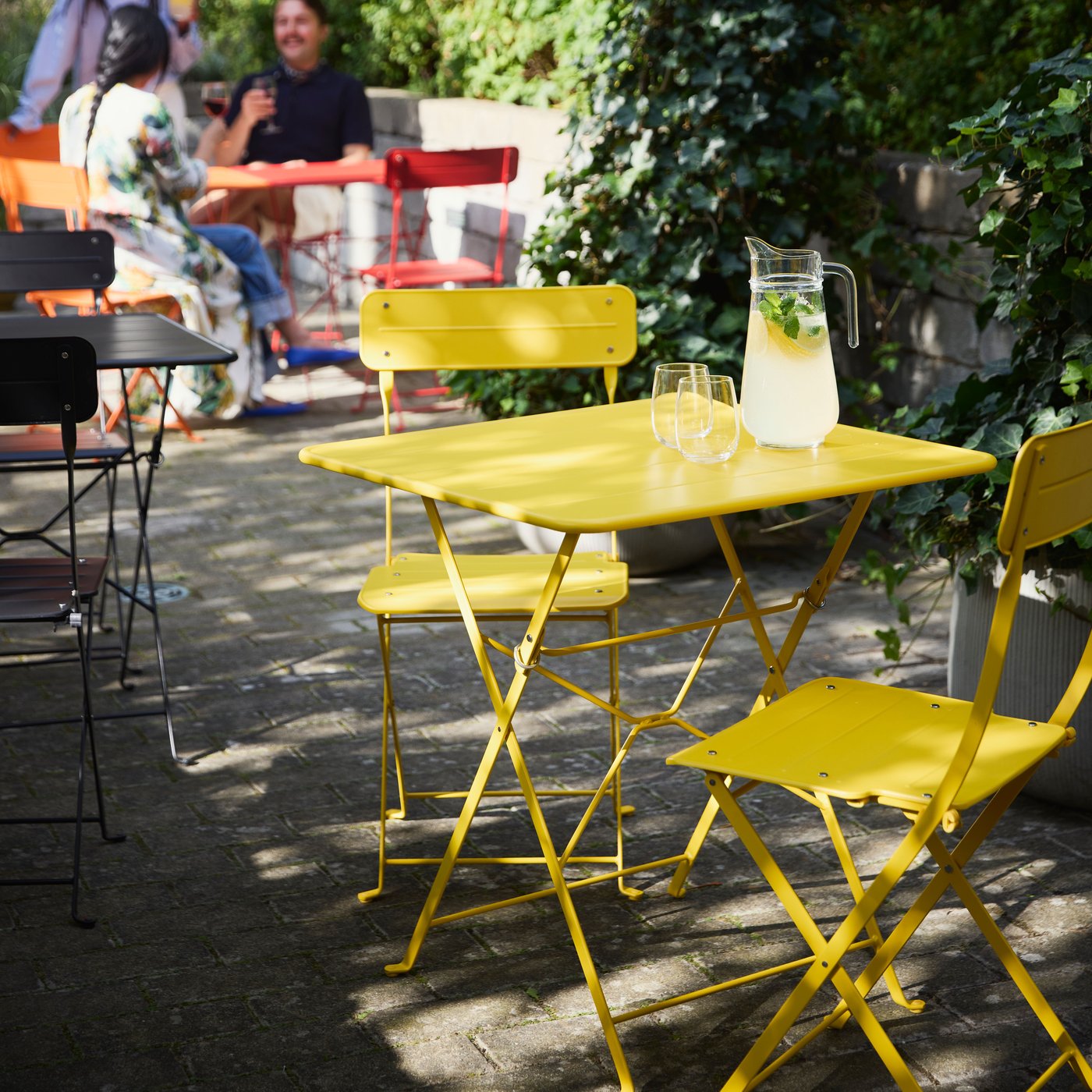 Bright SUNDSÖ tables in red, black, and yellow in a sunny courtyard, with people enjoying their time in the background.