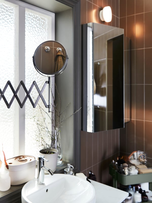 A mirror with an extension arm that’s pulled out over a wash-basin, a cabinet with mirror door and a grey-green trolley.