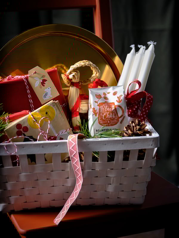 A decorated basket filled with a brass-coloured GLATTIS tray, EGENTID black chai tea and JUBLA candles.