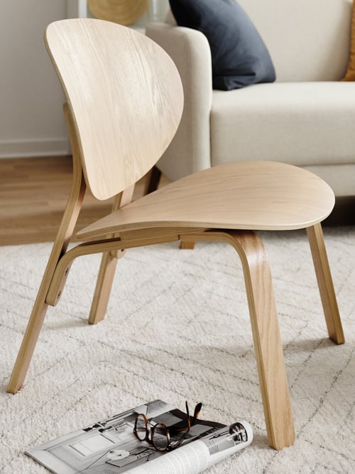 A light wood FRÖSET chair on a beige rug. A magazine is on the floor beside it with a pair of glasses on top.