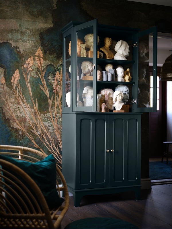 A dark green cabinet, the upper part of which has glass cabinet doors, in which various busts made of clay and stone are stored. 