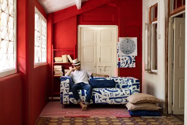 En barfot mann i panamahatt sitter i en blå og hvit sofa i et rom med røde vegger, ei bokhylle, ei hvit dør og et tofarget gulvteppe.