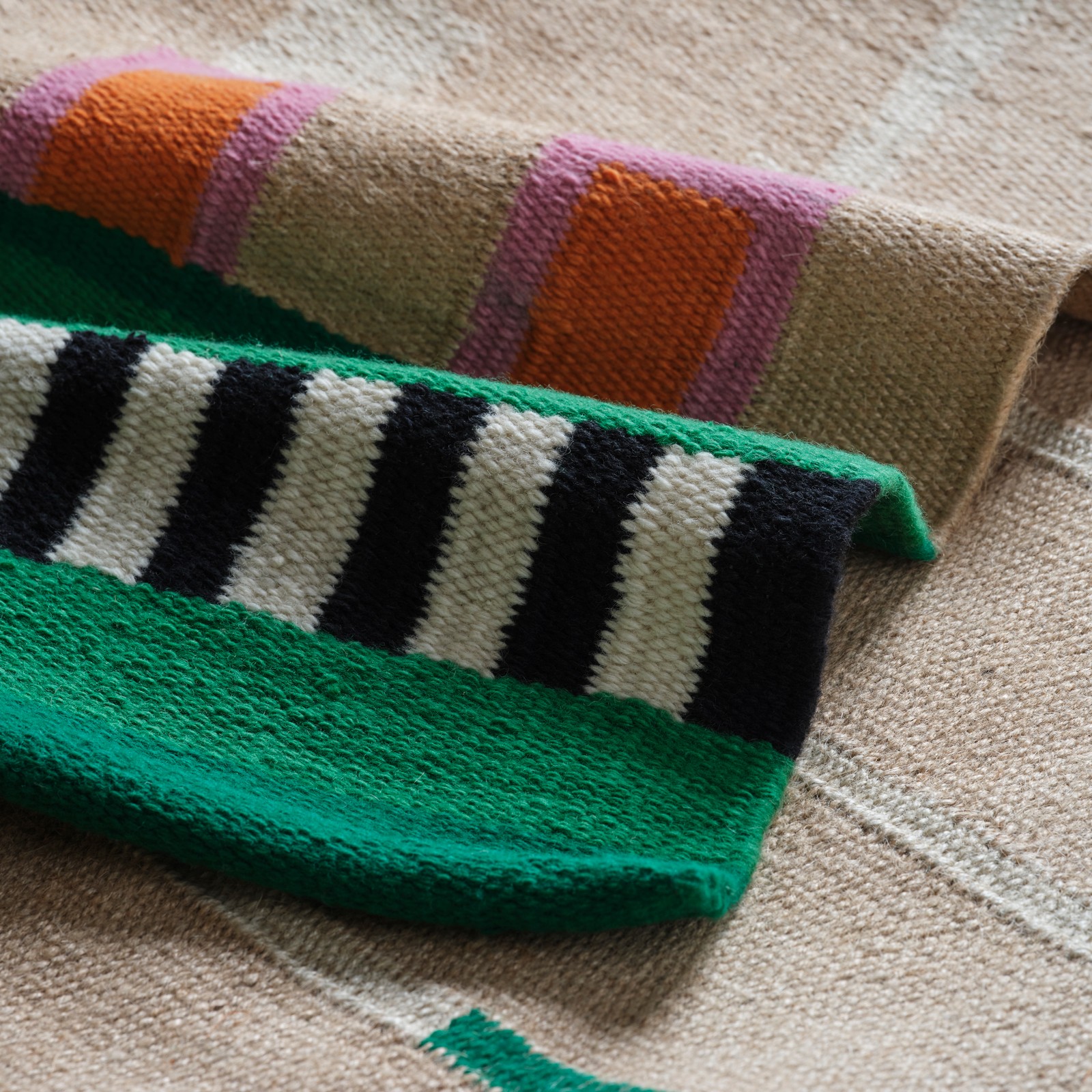 Un covor MÄVINN din iută nevopsită și bumbac, cu dungi albe și negre din lână vopsită, cu detalii verzi, roz și portocalii.