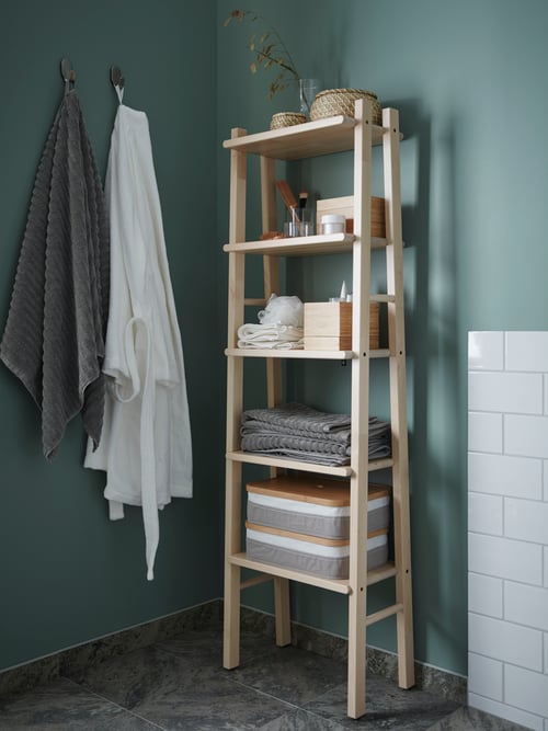 In einem grünen Badezimmer steht ein leiterähnliches VILTO Regal in Birke an einer Wand. In ihm befinden sich gefaltete Handtücher und verschiedene Behälter aus Seegras und Bambus.