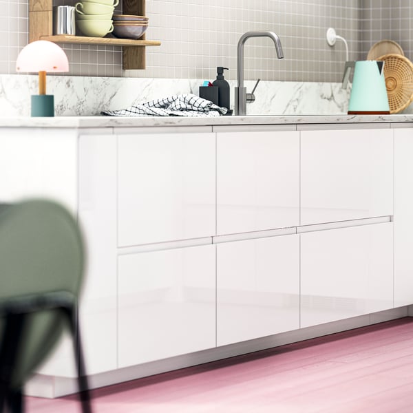 Frentes VOXTORP em branco brilhante, chão em rosa, cadeiras em verde e apontamentos de cor nas prateleiras numa cozinha moderna.