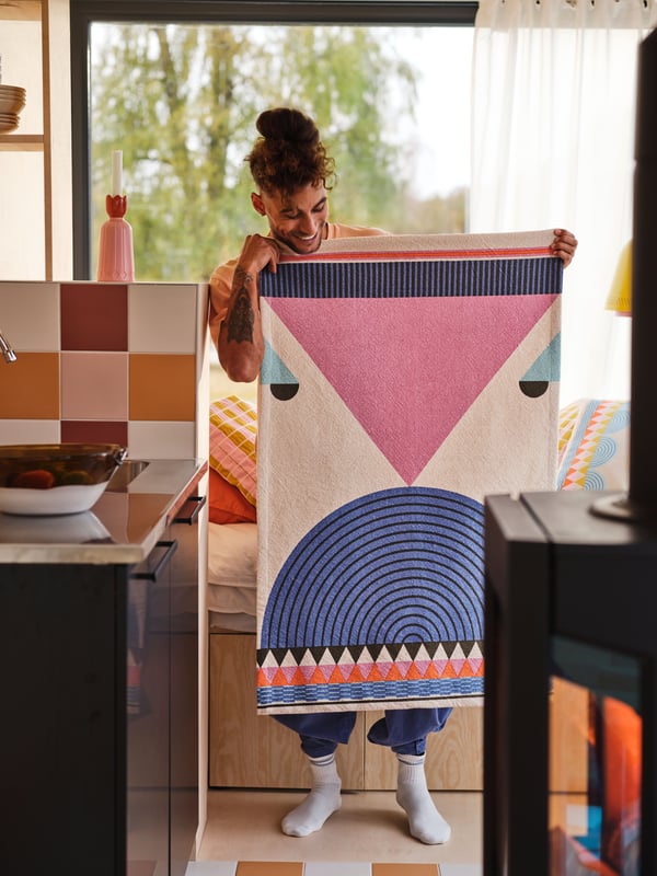 A person standing in a room holds an OMMJÄNGE hanging tapestry with both hands in front of themselves, smiling.
