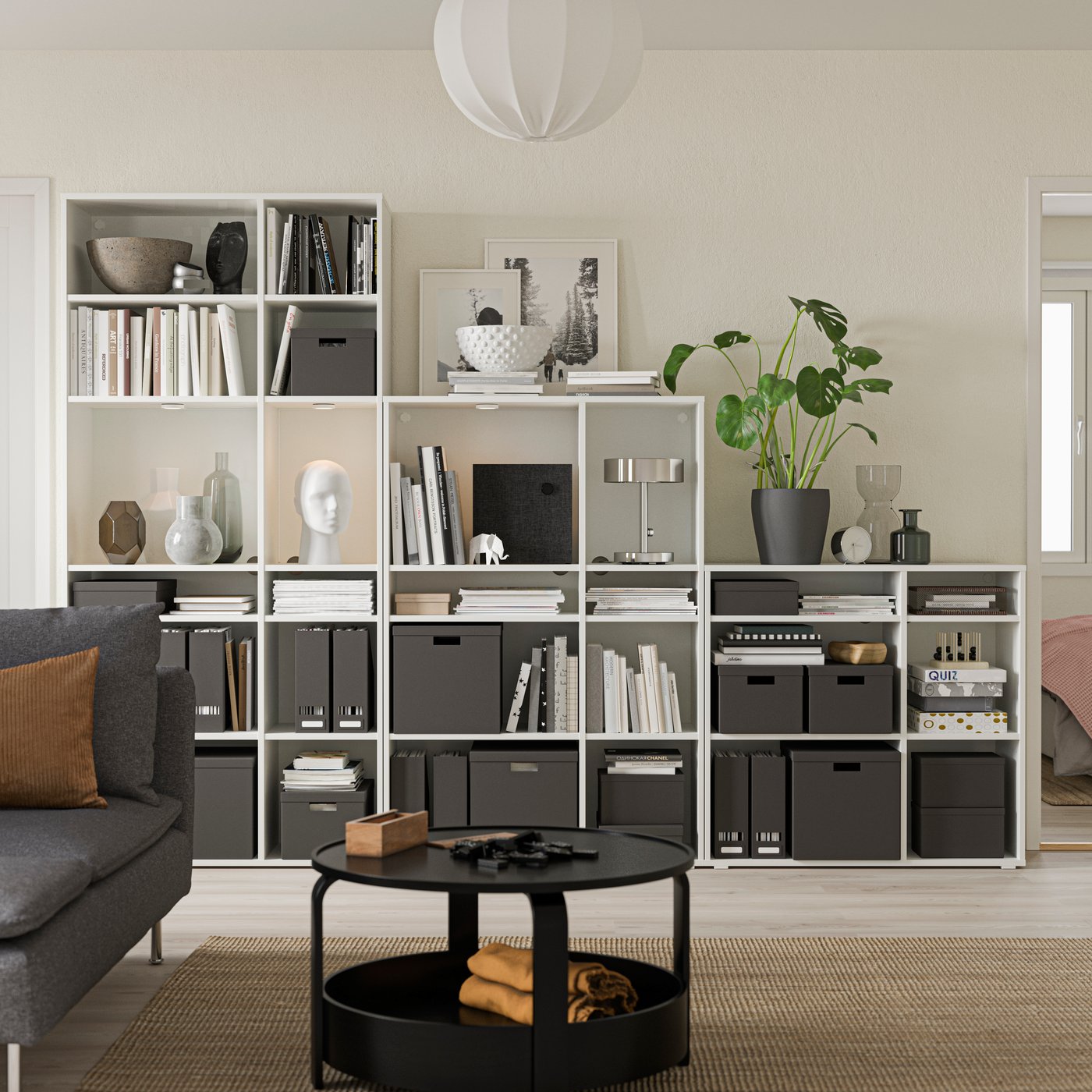 Vita öppna bokhyllor med svarta förvaringslådor och dekorativa föremål som vaser, böcker och skulpturer. En stor grön växt står på ett av hyllplanen. I förgrunden syns ett runt svart soffbord med en ljusbrun detalj, och till vänster en grå soffa med en brun kudde. Rummet har ljusa väggar och en rund taklampa i vitt.
