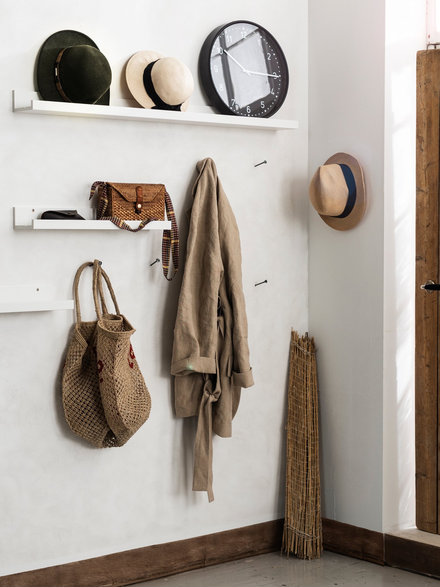 Entré med vita väggar och två hyllor med hattar, väska och accessoarer. Krokar med hängande korg och kläder, samt en trädörr med hatt på sidan.
