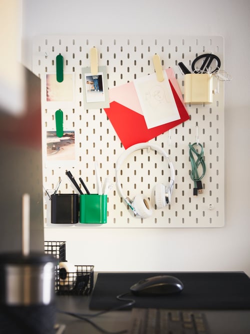 A white SKÅDIS pegboard on a wall by a desk holding stationery, notes, headphones and pots with pens and scissors inside.