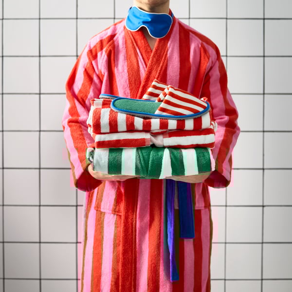 A man in a pink and red striped SKOGSSALLAT bathrobe holding a pile of green, white and red striped towels and slippers.