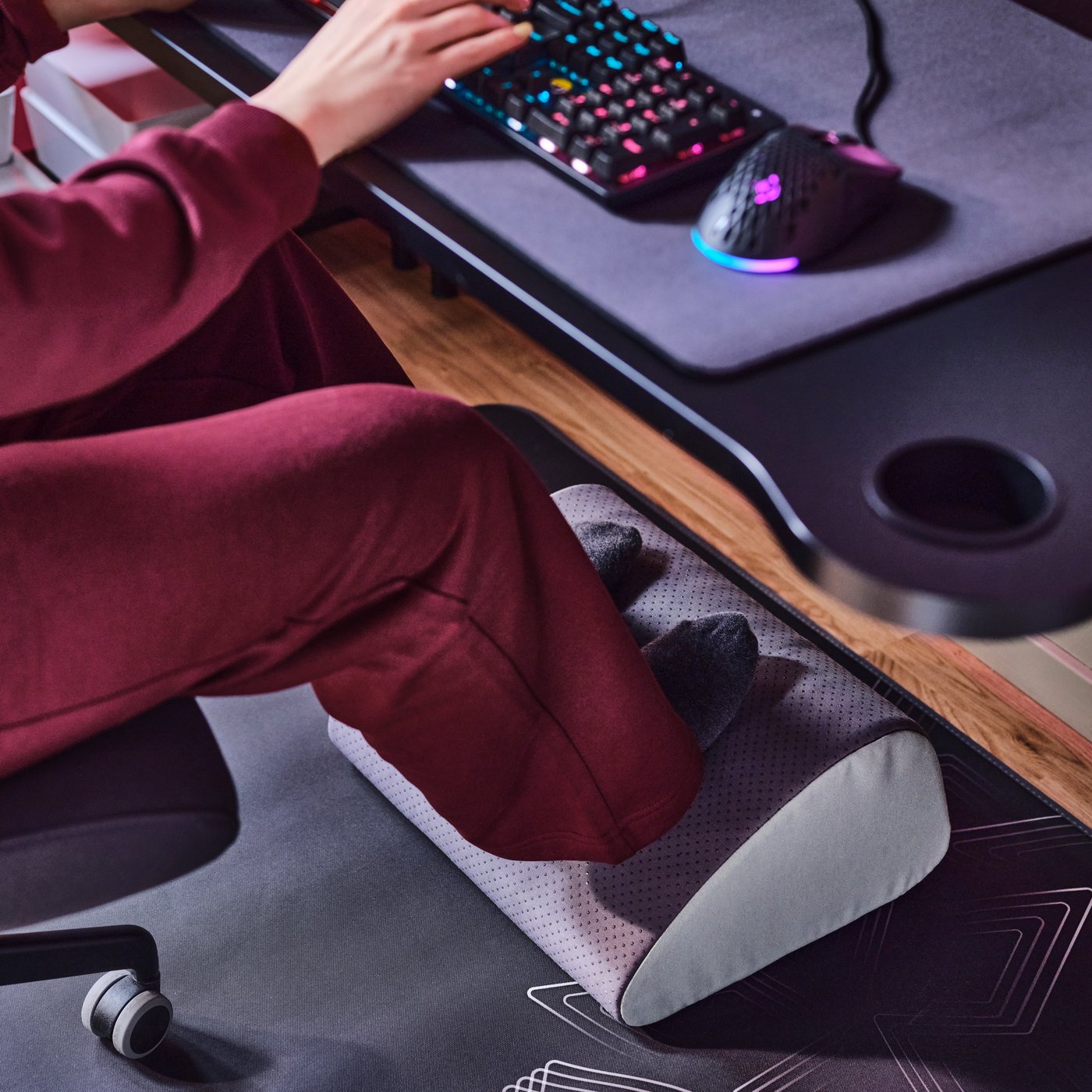 Una persona sentada en un escritorio FRÖSKELÅS con una estación de juego, con los pies apoyados en un reposapiés ergonómico multifuncional ÖVNING.