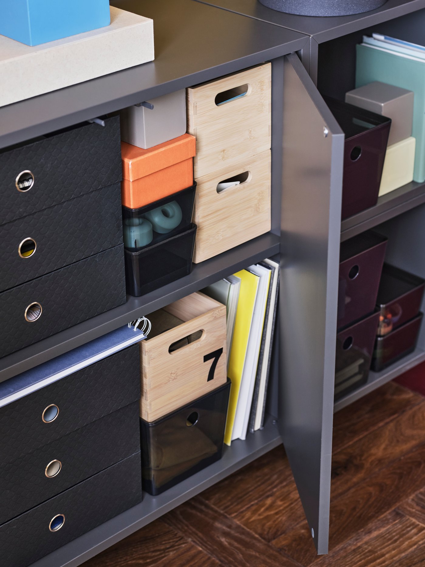A close-up of the inside shelves in a grey EKET wall cabinet organised with wooden and paper boxes in different sizes.