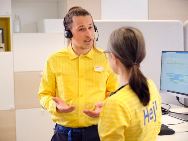 Två medarbetare i en kundservice- och kontorsmiljö står vid en arbetsstation och samtalar, med dator och headset som används för support och kundärenden.