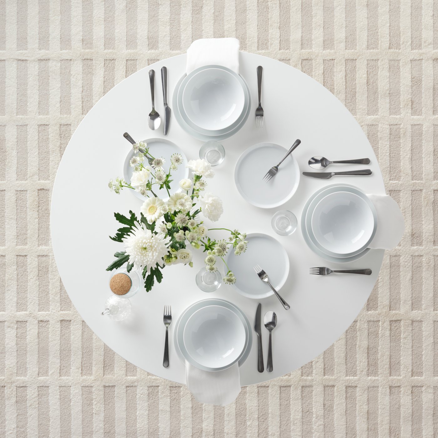 A round white table set for a meal with IKEA 365+ deep plates and side plates, a bunch of flowers and DRAGON cutlery.