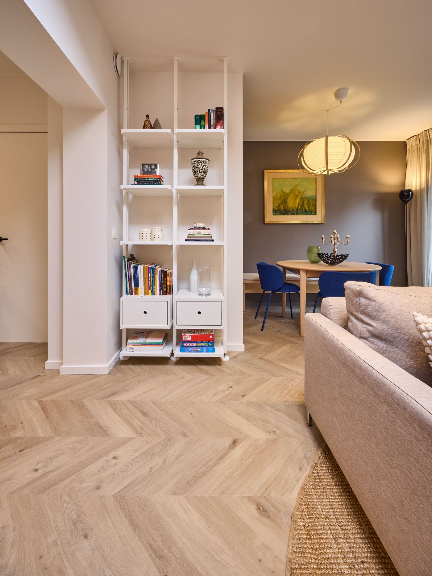 Woonkamer met witte kast vol boeken en decoratie, zicht op eettafel met blauwe stoelen, warme verlichting en lichte houten vloer.