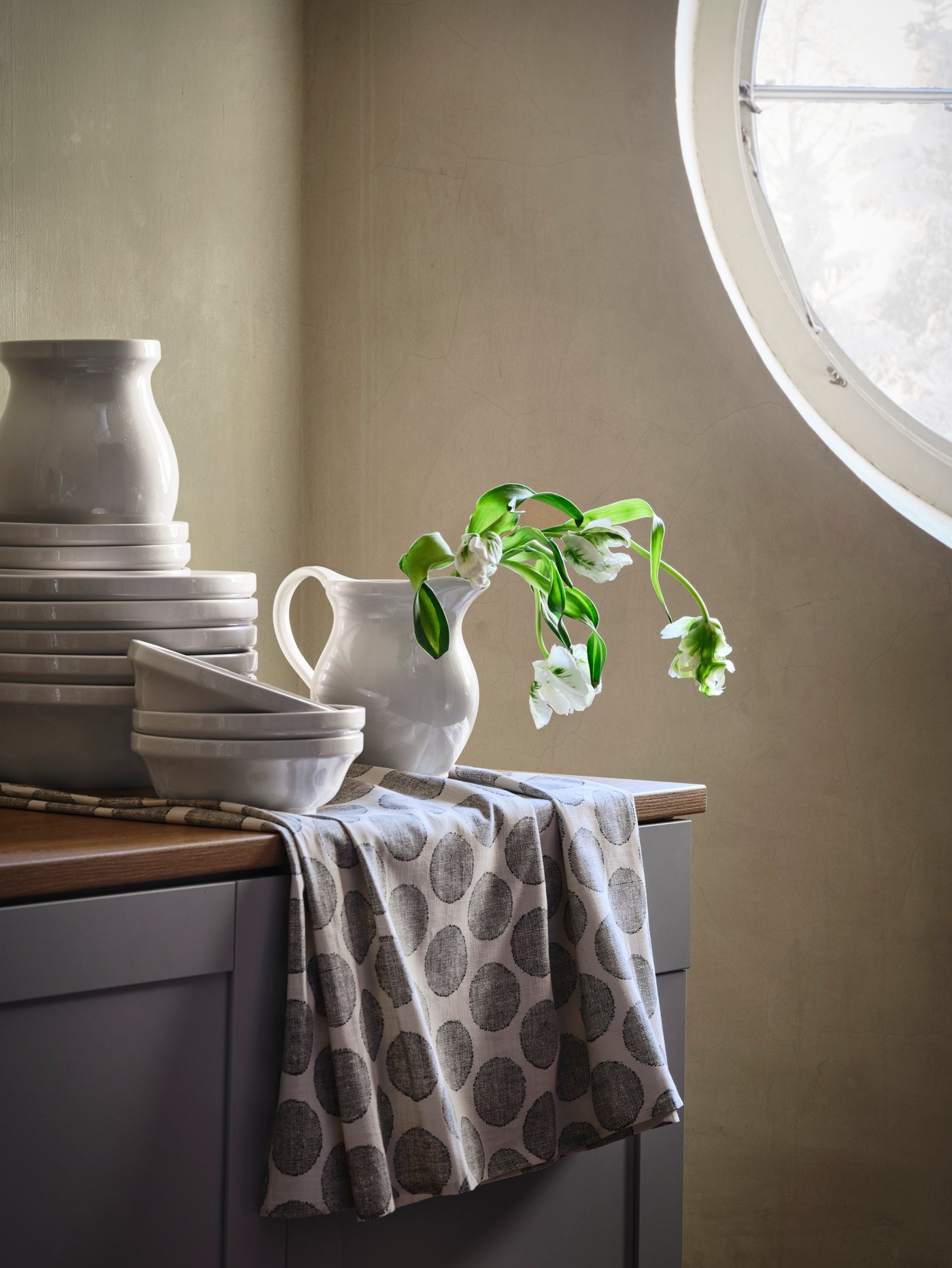 DEKORERA katilu, ontzi eta pitxer zuriak bata bestearen gainean jarrita sukaldeko ontzitegiaren gainean; pitxer batek loreak ditu.