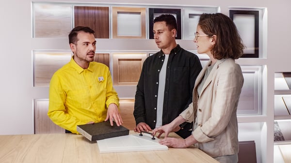 Konsultant IKEA stojący w żółtej koszulce przy stanowisku z frontami kuchennymi i próbkami blatów. Wskazuje na propozycję zestawu i prezentuje go parze klientów.