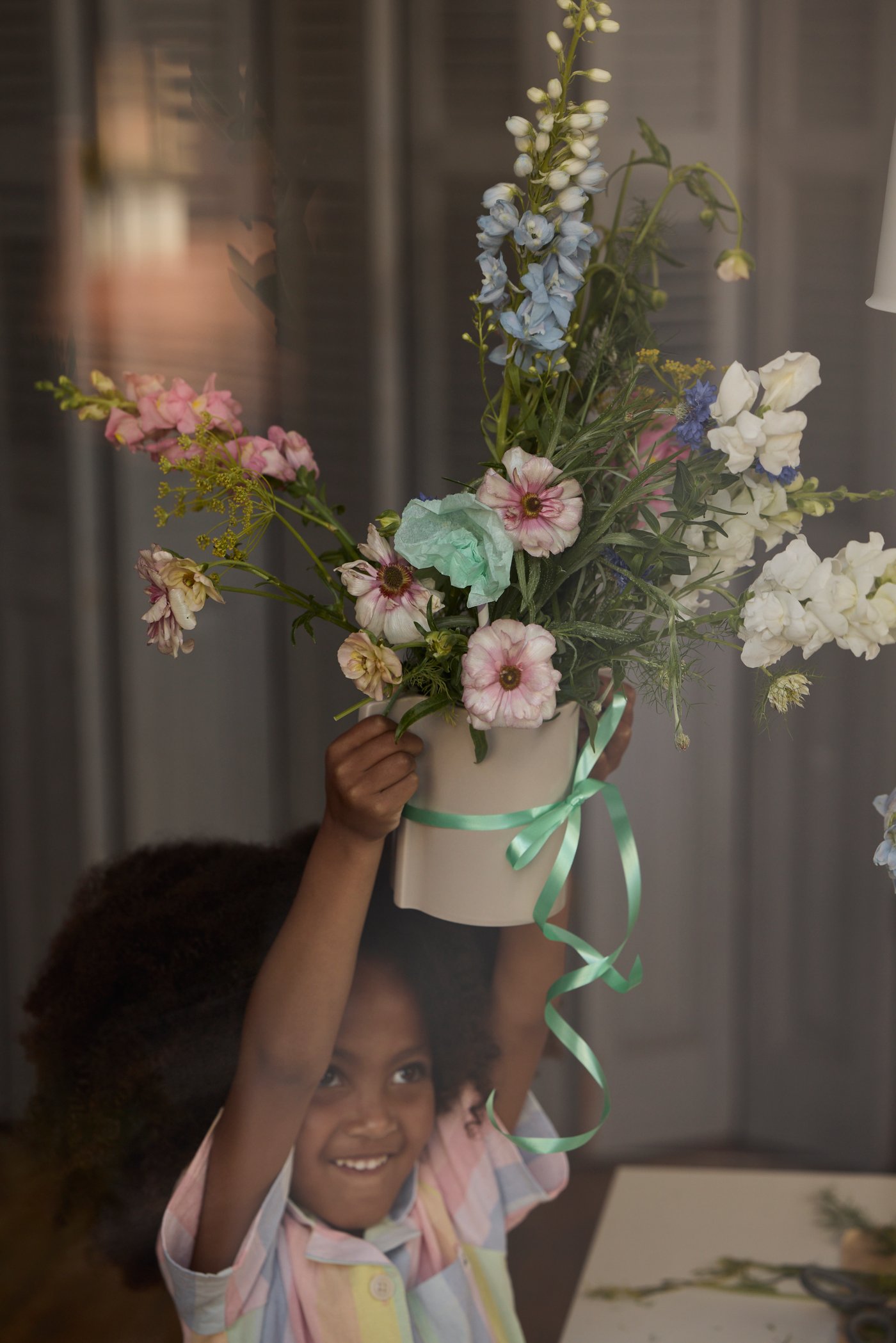 Ett leende barn håller upp en vacker bukett med färska vårblommor, med rosa, blå och vita blommor, i en kruka dekorerad med ett grönt band.