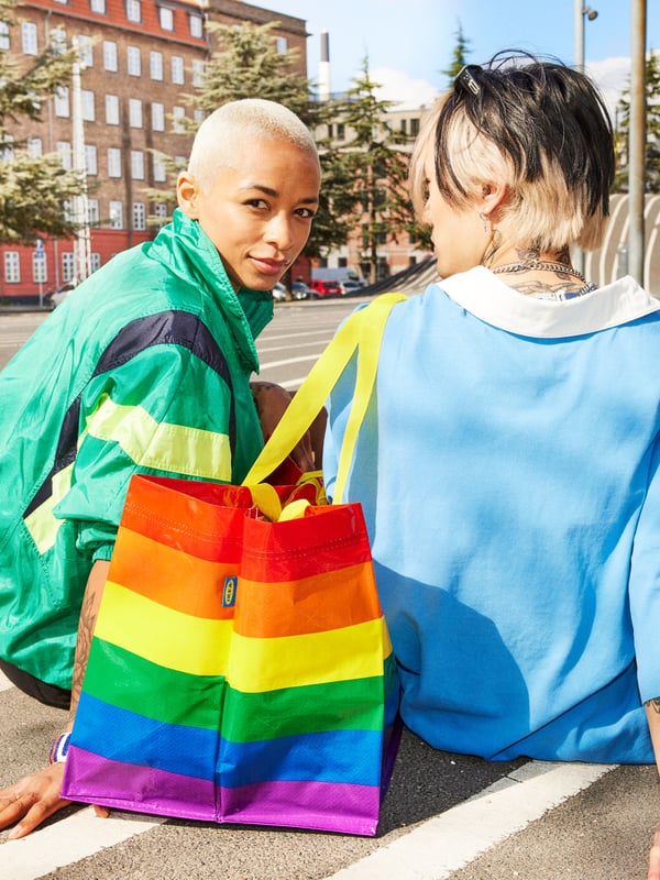 Two people hugging outside on a city street, one of them has a large STORSTOMMA bag, in rainbow colors, hanging on the shoulder. 