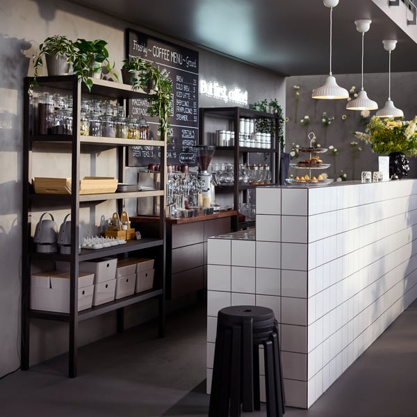 A café counter with dark brown STOCKHOLM 2025 shelving units in oak veneer/dark brown filled with glassware and jars.