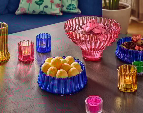 A dark wood table full of colourful glass AROMATISK tealight holders, vases and bowls