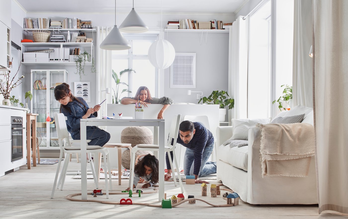 Un soggiorno bianco luminoso, dove una famiglia di quattro persone gioca con una pista costruita con i set di trenini in legno LILLABO.